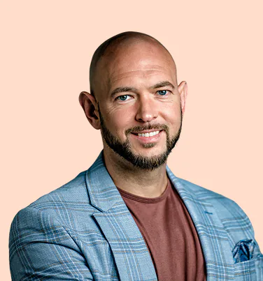 Smiling bald man with blue eyes and a beard wearing a blue checked blazer and a brown shirt against a peach background.